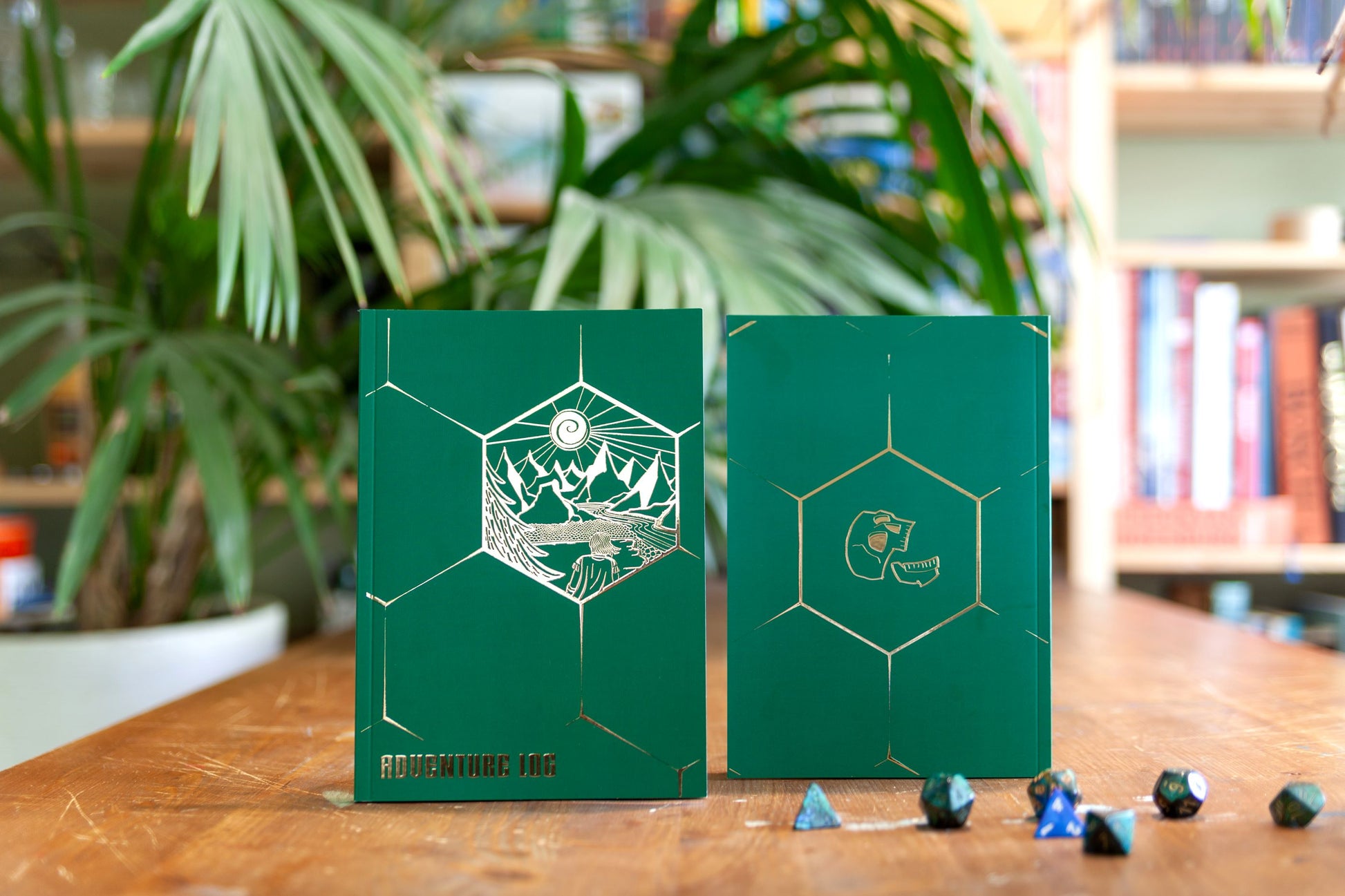 A notebook for Dungeons & Dragons 5E to write your stats and notes in. On the green cover is a gold foil illustration of an anonymous adventurer, staring at the mountains in the distance. The title, 'Adventure Log', is visible at the bottom left of the cover. On the back of the cover is a laughing skull: the logo of Cacklin Slayers.