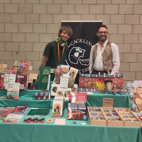 Marco and Josha standing behind a table filled with their products. The skull-logo of Cacklin Slayers is visible in the background.
